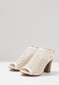 Bílé pletené sandály na podpatku s otevřenou špičkou, zadním páskem s přezkou a texturovaným hnědým dřevěným podpatkem.