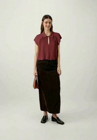 Plum pleated blouse with collar, keyhole detail, and cap sleeves paired with a long brown denim skirt. Model holds a brown handbag, wearing black loafers.