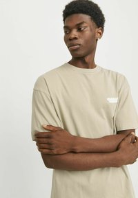 Beige bomulds T-shirt med korte ærmer, rund halsudskæring og et lille hvidt logo på venstre bryst. Glat tekstur, afslappet pasform.