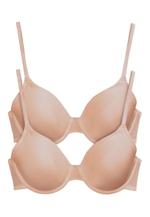 Femeie cu păr lung și închis la culoare, purtând un sutien bej și lenjerie de corp cu talie înaltă, așezată pe un perete gri deschis, privind către cameră.