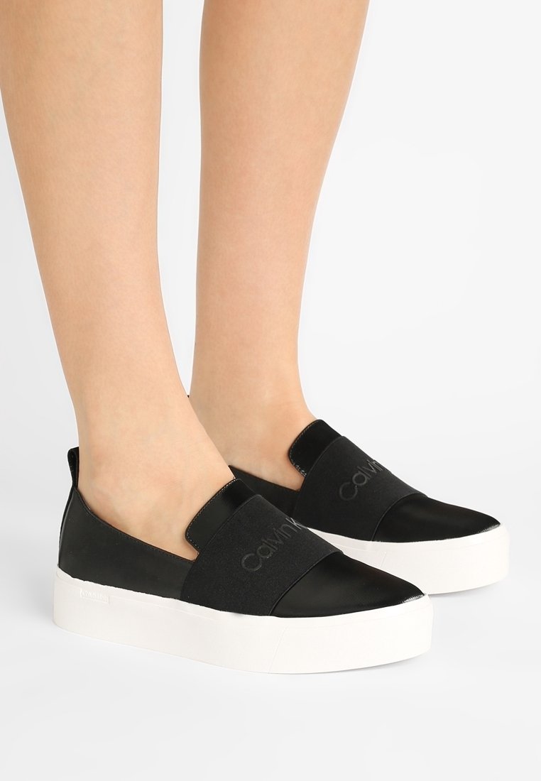 Jambes portant des baskets noires à enfiler avec des semelles blanches épaisses et de larges bandes élastiques sur le dessus, ornées du logo Calvin Klein.