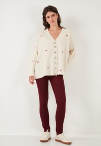 Cardigan in maglia crema con scollo a V, fiocchi ricamati e chiusura con bottoni grandi. Abbinato a pantaloni skinny bordeaux e sneakers bianche.