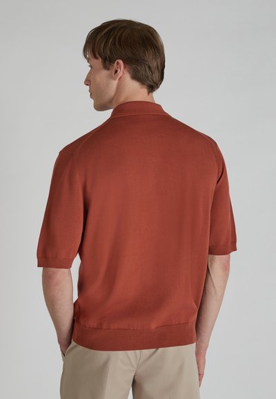 Polo à manches courtes en orange brûlé, texture tricotée, avec un col classique et un ourlet côtelé. Porté avec un pantalon kaki.