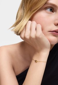 Bracciale d'oro con una delicata catena e un piccolo ciondolo, indossato su un polso. Lo sfondo è neutro, evidenziando il design e il materiale del gioiello.