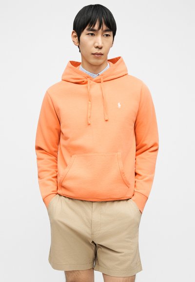 Νέος άνδρας που φοράει φωτεινή πορτοκαλί φούτερ με μπροστινή τσέπη και μπεζ σορτς, στέκεται με τα χέρια στις τσέπες μπροστά σε απλό φόντο.