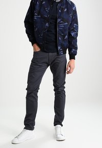 Chaqueta bomber de la Marina con patrón abstracto, detalles en negro, combinada con pantalones oscuros ajustados de color gris y zapatillas blancas. Texturas suaves y diseño moderno.