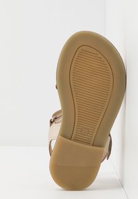 Dětské béžové sandály s gumovou podrážkou zespodu, velikost 25 označena na podrážce, umístěné u bílých rohových stěn.