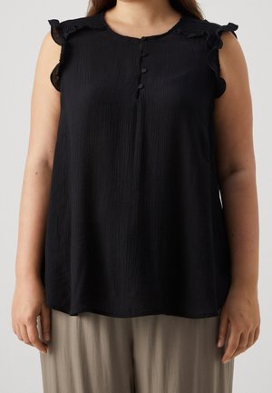 Femme portant un débardeur noir sans manches avec des épaules volantées et trois boutons au col, associé à un pantalon beige ample.