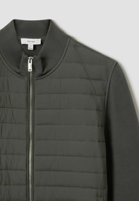 Veste bomber vert olive avec fermeture éclair à l'avant, présentant une texture matelassée sur le corps et des manches en tissu lisse. Col et poignets côtelés.