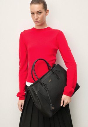 Piros pulóveres és fekete szoknyás nő, aki nagy fekete bőr kézitáskát tart, dupla fogantyúval és arany logóval.