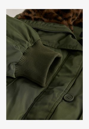 Giacca isolante verde con polsini a coste, chiusura con zip e collo in pelliccia marrone a motivo leopardato. Include un dettaglio con bottone nero.