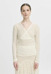 Abito in pizzo crema con un design avvolgente, maniche lunghe e delicate finiture a scallop. Il motivo floreale su tutto l'abito arricchisce la texture e l'interesse visivo.