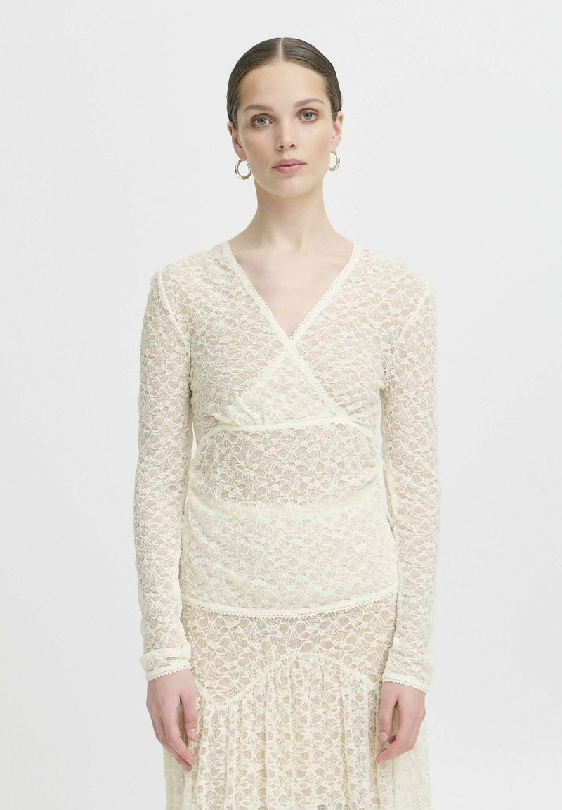 Abito in pizzo crema con un design avvolgente, maniche lunghe e delicate finiture a scallop. Il motivo floreale su tutto l'abito arricchisce la texture e l'interesse visivo.