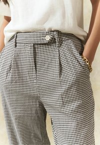 Un pantalon à carreaux noir et blanc avec une taille haute, des plis et une fermeture à bouton à l'avant. Porté avec un haut de couleur claire et un bracelet en or.
