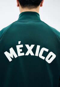 Persoon met een donkergroene jas met een hoge kraag, witte hoofdletters "MÉXICO" in boogvorm over de bovenrug.