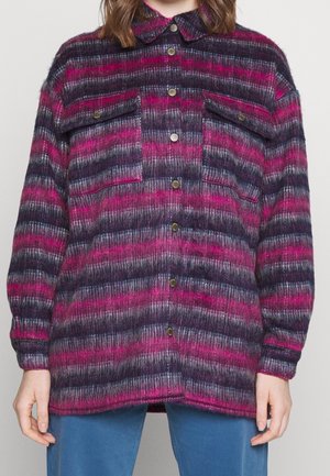 Kobieta ubrana w zapinaną na guziki, długorękawową koszulę-ramoneskę z kołnierzykiem w poziome paski w kolorach różowym, fioletowym i czarnym, w połączeniu z niebieskimi spodniami.