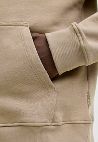 Ręka o ciemnej karnacji w beżowej kieszeni swetra z widocznym ściągaczem i metką "CORE" na bocznej szwie.