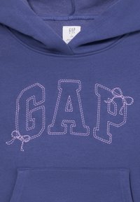 Modra otroška kapuca s pisano oznako "GAP" v velikih vezenih črkah, pri čemer sta črki "G" in "P" okrašeni z majhnimi vezenimi pentljami.