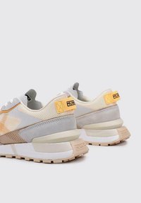 Zapatillas con parte superior de malla en gris claro y beige, acentos amarillos y paneles texturizados. Suela gruesa en blanco y crema con un detalle del logo en la parte trasera.