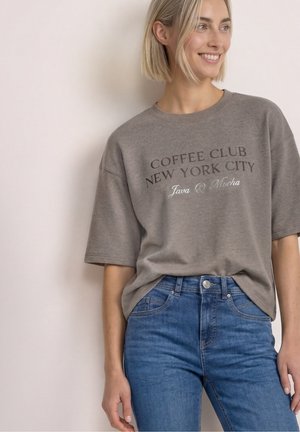 Donna sorridente che indossa una maglietta grigia oversize con il testo "Coffee Club New York City Java & Mocha" e jeans blu, in piedi contro un muro semplice.