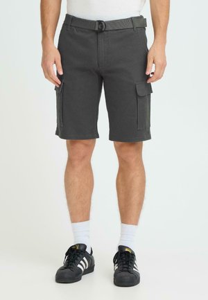 Mann trägt dunkelgraue Cargoshorts mit Klappentaschen, weiße Socken und schwarze Adidas-Sneaker mit weißen Streifen, steht vor weißem Hintergrund.