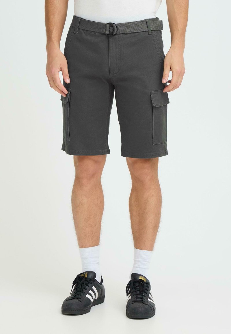 Homme portant un short cargo gris foncé avec poches à rabat, des chaussettes blanches et des baskets Adidas noires à rayures blanches, debout devant un fond blanc.