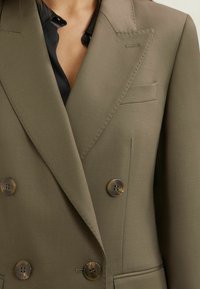 Primo piano di una persona che indossa un blazer doppiopetto verde oliva con bottoni marroni sopra una camicetta di seta nera.