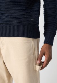 Navy strikkegenser med teksturert søm og ribbestrikkede mansjetter, kombinert med lysebeige bukser med sidelommer.
