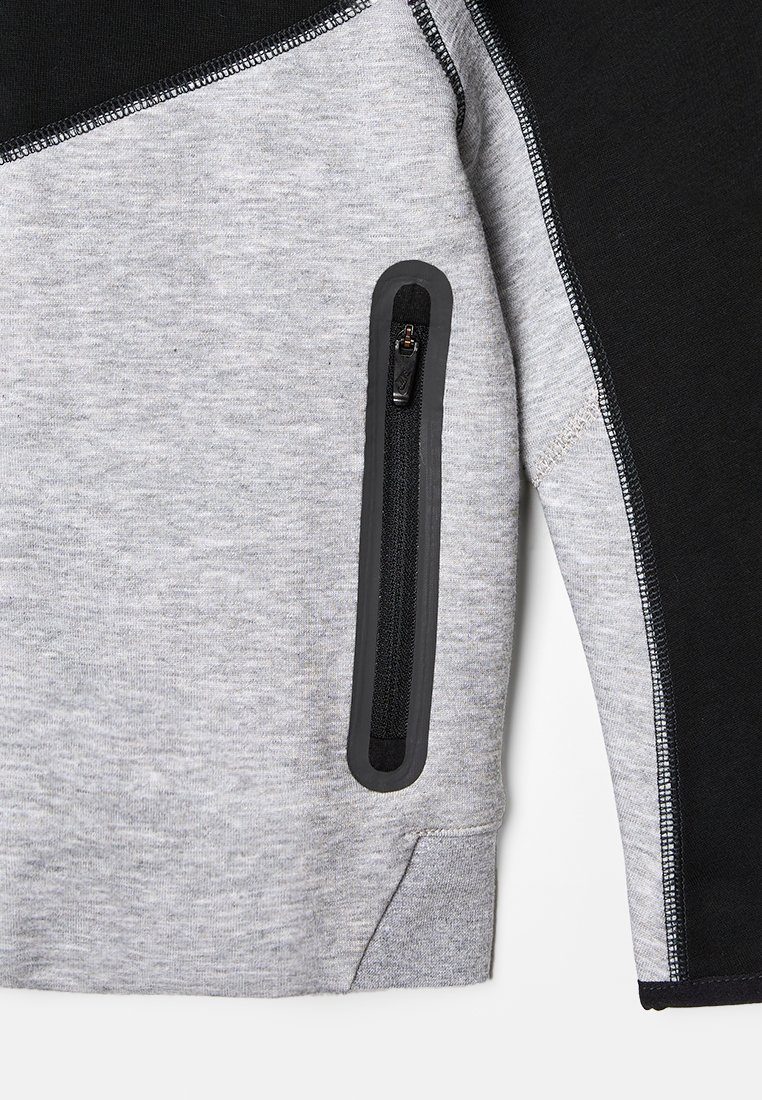 Grijze en zwarte sweatshirt met een ritszak, gestructureerde stof, contrasterende panelen en zichtbare stiksels langs de naden.