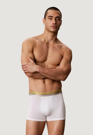 Umięśniony mężczyzna z krótkimi włosami stoi bez koszulki z założonymi na piersi rękami, nosząc białe bokserki Calvin Klein ze złotym paskiem w pasie.