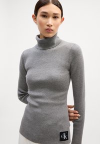 Grau gerippter Rollkragenpullover mit langenÄrmeln und taillierter Form; verfügt über ein schwarzes CK-Logo-Patch am Saum. Weicher Stoff und dehnbares Material.