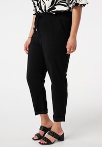 Pantalons noirs à taille élastique avec poches latérales, forme fuselée et poignets retroussés, associés à des sandales à talons blocs ouvertes et rembourrées noires.
