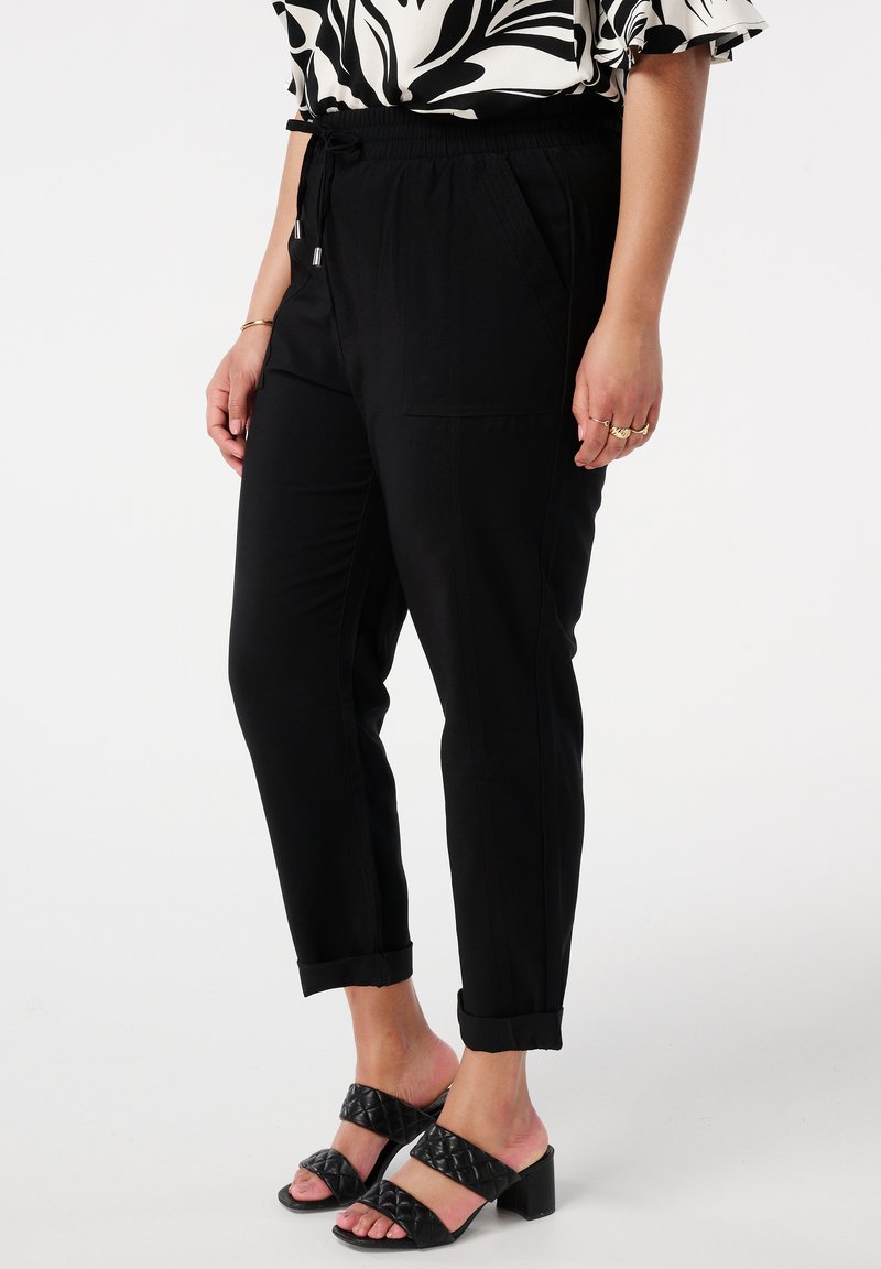 Pantalons noirs à taille élastique avec poches latérales, forme fuselée et poignets retroussés, associés à des sandales à talons blocs ouvertes et rembourrées noires.