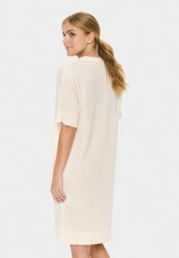 Femme aux longs cheveux blonds attachés en queue de cheval, portant une robe ample couleur crème, arrivant au genou et à manches courtes, se tenant debout et se retournant par-dessus son épaule.