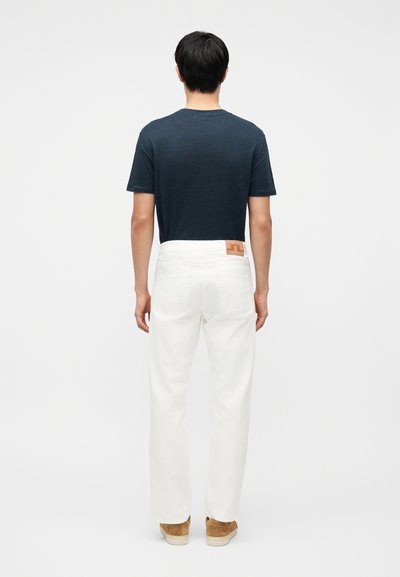 Άνδρας που φοράει σκούρο μπλε μπλουζάκι, λευκό τζιν με δερμάτινο πατζάκι στη μέση, και ανοιχτό καφέ παπούτσια, στέκεται με την πλάτη προς τα εμάς.