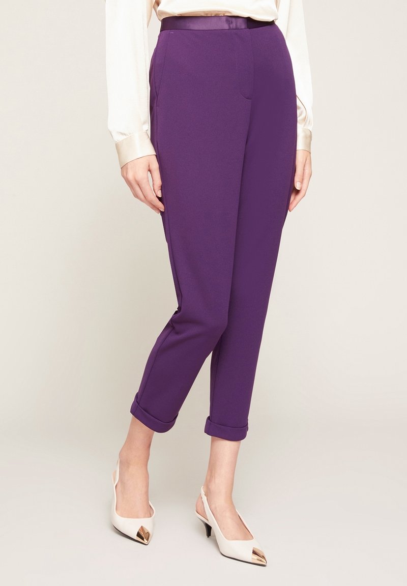 Pantalon violet fuselé avec une texture lisse, des revers roulés et une ceinture élégante. Porté avec des talons blancs ornés d'un accent métallique.