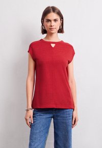 Top rosso a maniche corte con dettaglio alla scollatura a chiave. Realizzato in un tessuto leggero con una texture morbida, abbinato a jeans di denim blu.