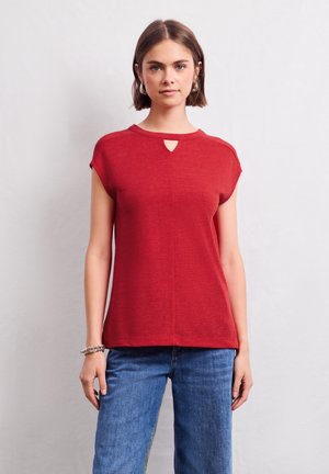 Haut rouge à manches courtes avec un détail d'encolure en œil de clé. Fabriqué dans un tissu léger avec une texture douce, associé à un jean en denim bleu.