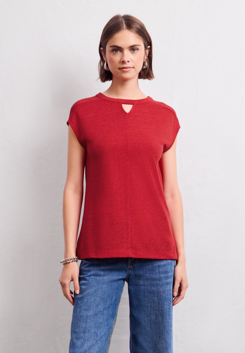 Top rosso a maniche corte con dettaglio alla scollatura a chiave. Realizzato in un tessuto leggero con una texture morbida, abbinato a jeans di denim blu.