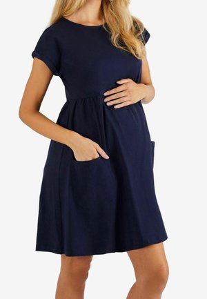 Femme portant une robe de grossesse bleu marine à manches courtes, une main sur le ventre et l'autre dans une poche avant.