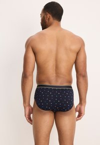 Slip da bagno da uomo in navy con un motivo a triangoli verdi e blu. Presenta una fascia elastica gialla in vita. Il materiale sembra liscio ed elastico.