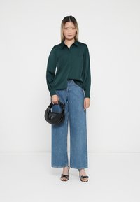 Mørkegrønn, langermet skjorte med knapprekkefront; kombinert med vide, lyseblå jeans; holder en svart, buet håndveske; iført svart og hvit sandaler.