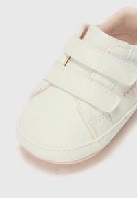 Chaussure en simili cuir blanc avec deux bandes Velcro, accents roses, bout arrondi et détails de couture. Doublure intérieure douce pour le confort.