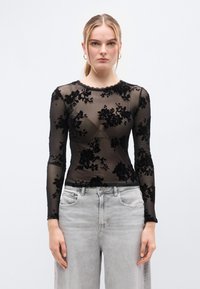 Μαύρο διάφανο μακρυμάνικο τοπ με βελούδινο λουλουδάτο σχέδιο, ντεκολτέ με κυματιστό τελείωμα και στενή γραμμή, συνδυασμένο με ανοιχτό γκρι φαρδιά τζιν παντελόνια.