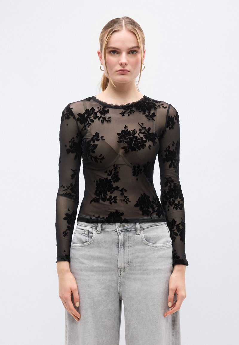 Μαύρο διάφανο μακρυμάνικο τοπ με βελούδινο λουλουδάτο σχέδιο, ντεκολτέ με κυματιστό τελείωμα και στενή γραμμή, συνδυασμένο με ανοιχτό γκρι φαρδιά τζιν παντελόνια.