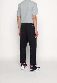 Personne portant un t-shirt gris à manches courtes, un pantalon noir et des baskets blanches, noires et rouges, debout de dos devant un fond uni.