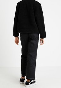 Zwarte fleecejack met een gestructureerd oppervlak, gecombineerd met rechte zwarte broek en zwarte sneakers met witte accenten. Zichtbare stiksels.