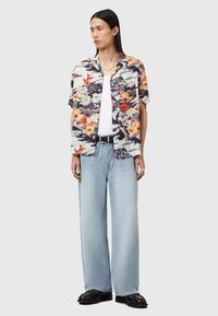 Camicia a maniche corte con motivi floreali in nero, bianco, arancione e blu, abbinata a jeans larghi blu chiaro e scarpe nere.