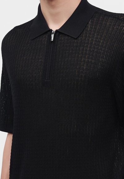 Homme portant un polo en maille noire à manches courtes avec un motif texturé et une fermeture éclair devant.
