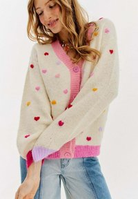 Dzianinowy kardigan w kremowym kolorze z kolorowymi wzorami serduszek, różowe zapięcie na guziki, kontrastowe różowe wykończenia przy dekolcie i mankietach, oraz luźny krój.